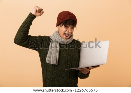 Portrait of a happy young african man dressed in autumn clothes isolated over beige background, holding laptop computer, celebrating success