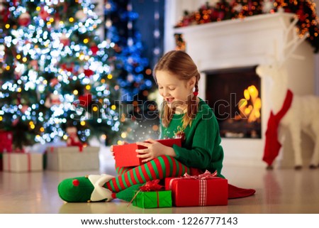 Child opening present at Christmas tree at home. Kid in elf costume with Xmas gifts and toys. Little girl with gift box and candy at fireplace. Family celebrating winter holidays. Home decoration.