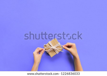 Woman hands give wrapped valentine or other holiday handmade present in paper with pink ribbon. Present box, decoration of gift on purple table, top view with copy space.