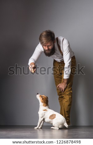 A man with a beard in a white shirt and yellow suspenders playing with his pet dog Jack Russell Terrier on a gray background
