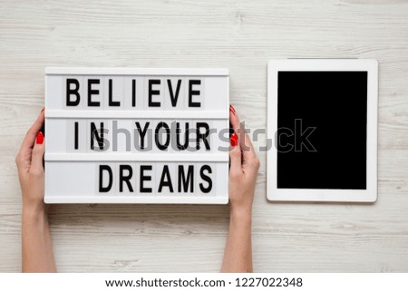 Female hands hold modern board with text 'Believe in your dreams', tablet wit blank screen over white wooden background. From above, overhead, flat-lay.