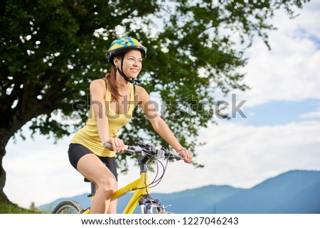 Attractive happy woman cyclist riding on yellow mountain bike under big tree, wearing helmet, enjoying summer day in the mountains. Outdoor sport activity, lifestyle concept