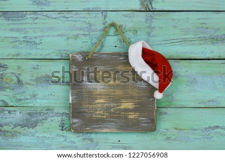 Blank wood sign with red and white Christmas Santa Claus hat hanging on antique rustic teal blue wooden door; holiday background with painted copy space