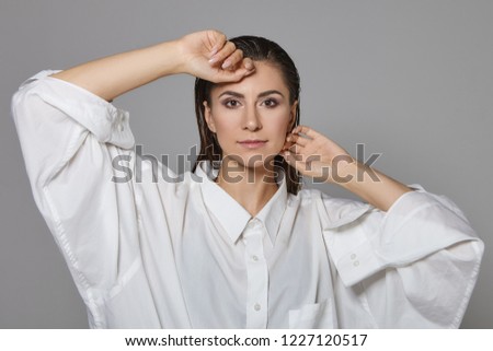Style and fashion concept. Picture of beautiful glamorous brunette female model with smoky eyes make up and dark hair combed back posing isolated wearing oversize white shirt, hands at her face