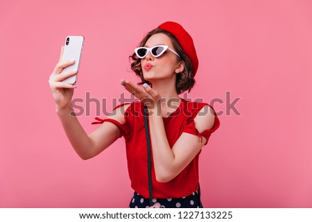 Stunning french woman sending air kiss while taking picture of herself. Indoor portrait of romantic curly lady in betet making selfie on pink background.