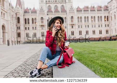 Emotional girl with gorgeous wavy hair talking on phone and drinking coffee on architecture background. Happy caucasian woman in retro casual attire looking around with smile.