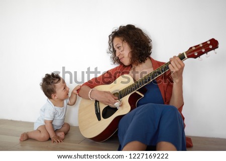 Picture of cute barefooted one year old infant boy sitting on floor with his positive young mom who is playing guitar, singing his favorite lullaby. People, music, children and entertainment
