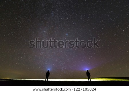 Back view of two men with head flashlights on green grassy field under beautiful dark blue summer starry sky. Night photography, beauty of nature concept. Wide angle shot, copy space background.