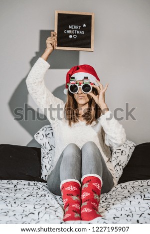 
Young and cheerful woman sitting on her bed at Christmas time with a blackboard that says "Merry Christmas". Wearing comfortable clothes for winter. Lifestyle.