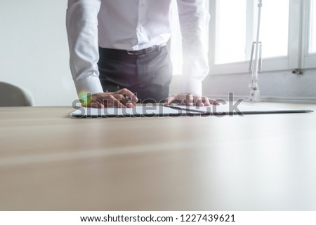 Businessman standing at his office desk leaning to sign a contract or document in a folder with lens flare and copy space.