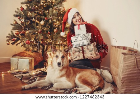 happy girl in santa hat holding gift boxes and cute dog sitting at golden beautiful christmas tree with lights and presents in festive room. family happy moments. winter  holidays