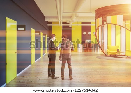 Businessmen talking in office reception hall with gray and yellow walls, row of yellow doors, reception desk and lounge area with computer. Toned image