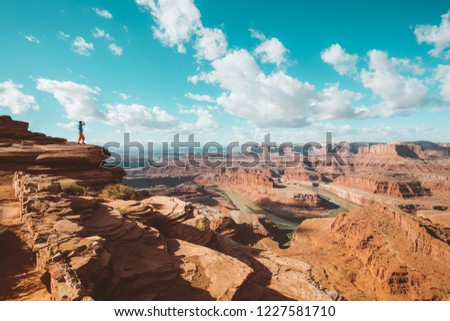A young male hiker is standing on the edge of a cliff enjoying a dramatic overlook of the famous Colorado River and beautiful Canyonlands National Park in scenic Dead Horse Point State Park, Utah, USA Royalty-Free Stock Photo #1227581710