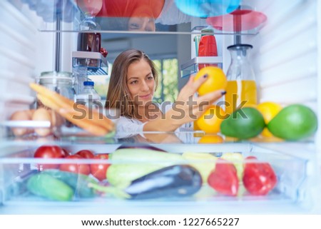 Woman standing in front of opened fridge and taking orange fruit. Fridge full of groceries. Picture taken from the iside of fridge.