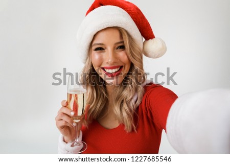 Portrait of a smiling blonde woman dressed in red New Year costume standing isolated over white background, holding glass of champagne, celebrating, taking a selfie