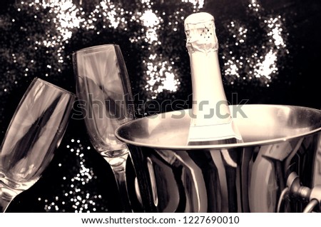 Champagne bottle in ice bucket and two tall glasses on blurred fireworks background. Close up on drink for toast and anniversary celebration. Black and white image 