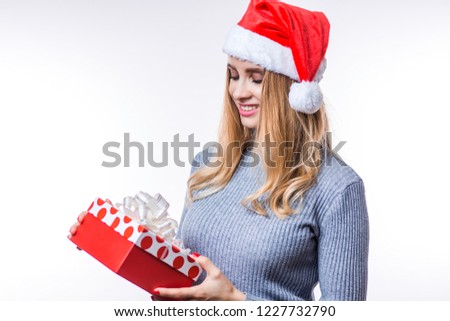 Happy pretty young woman holding gift box over white background. Beautiful girl wearing a gray sweater and a red Santa hat