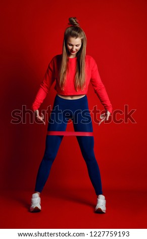 Woman with good physique doing stretching work out with elastic bands. Sports girl in fashionable sportswear on red background. Strength and motivation.