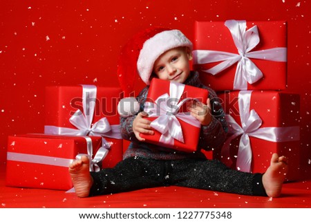 Smiling funny child boy in Santa red hat holding Christmas gift in hand and looking away on red background. Christmas concept. snowflakes on bokeh background