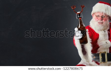 Santa Claus raising a Christmas toast with a bottle of beer decorated with deer antlers over grey with copy space