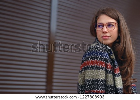 Thoughtful young woman wearing glasses and a tweed coat posing on brown urban background