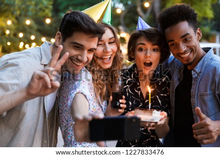 Group of excited multhiethnic friends celebrating with a cake outdoors, taking a selfie