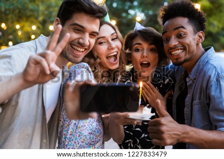 Group of joyful multhiethnic friends celebrating with a cake outdoors, taking a selfie