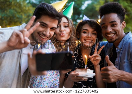 Group of smiling multhiethnic friends celebrating with a cake outdoors, taking a selfie