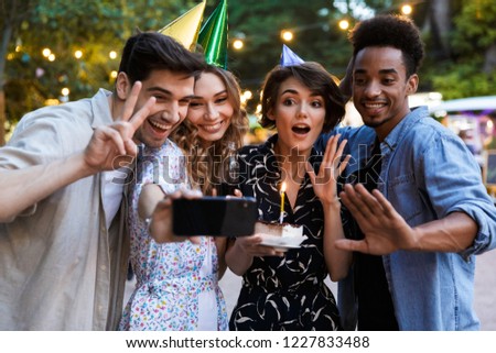 Group of happy multhiethnic friends celebrating with a cake outdoors, taking a selfie