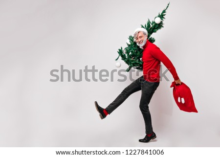 Full-length portrait of dancing santa claus with bag full of presents. Indoor shot of laughing grandpa with christmas tree.