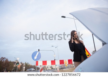 A young, beautiful businesswoman looking under the hood of a broken car and talking on the phone.