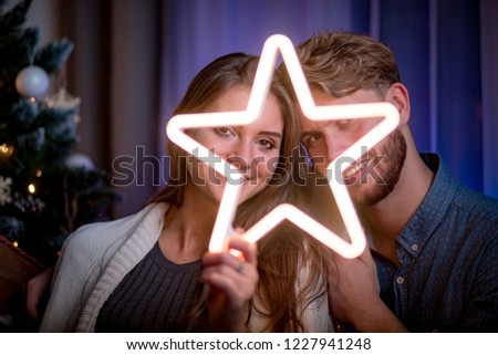 Cheerful couple sitting next to christmas tree and holding glowing LED star in dark cozy room at xmas eve