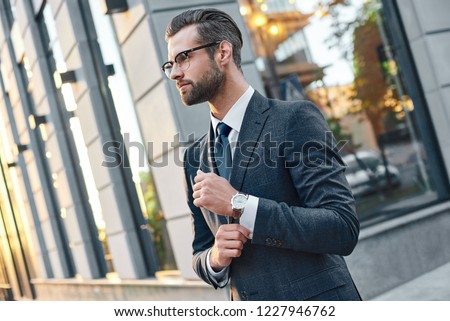 Close up portrait of a successful young bearded guy in suit and glasses. So stylish and nerdy. Outdoors on a sunny street, fixing his cuffs