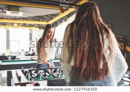 Two beautiful twin girls play table football and have fun. One of the sisters holds a toy ball in his hand and shows the tongue.