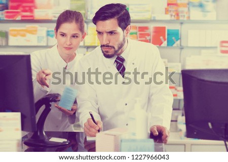 Two serious positive pharmacists are attentively stocktaking medicines with computer and note on paper in pharmacy.