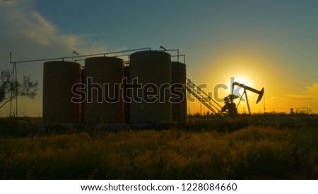 CLOSE UP: Industrial oil pump jack pumping crude oil and storing it in big reservoir containers in oil field. Nodding donkey pump working over the setting sun in the middle of desert at golden sunset