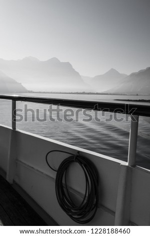 Black and white picture of the railing of a boat sailing on a foggy lake between the mountains