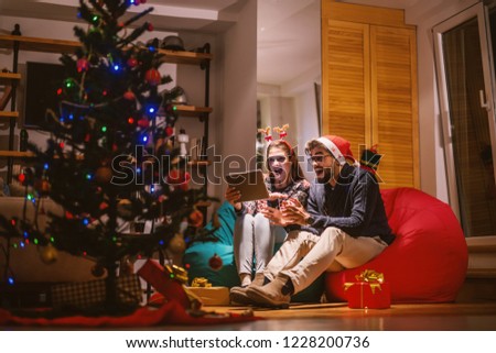 Couple using tablet while sitting in bean bags. On heads reindeer headband and santa's hat. In foreground Christmas tree and presents. Christmas holidays concept.