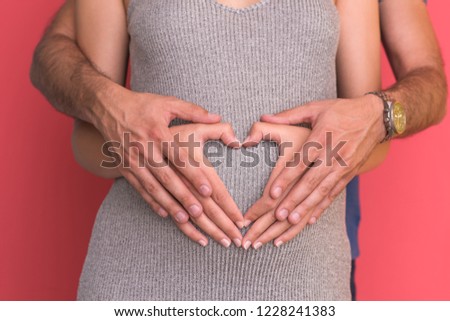 Happy young couple showing heart with their hands on belly of pregnant wife isolated on red background