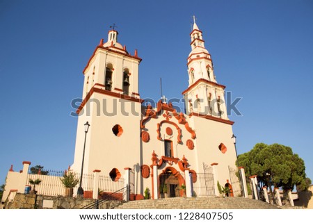 Full side shot of the front façade of a town traditional colonial church in Santiago, Nuevo León, México. Cream and orange contraste. Tree in a side.