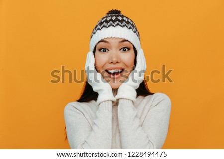 Woman portrait. Accessories. Warmness. Happiness. Asian girl in a white polo neck, cap and gloves is touching her face and smiling, on an orange background