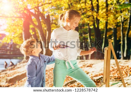 Children drawing with colorful paints outdoor in autumn park. Creative brother and sister painting on nature. Open air activity for different age children concept.