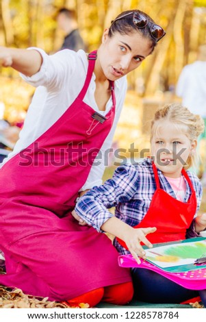Young female teacher and happy girl in red aprons are painting outdoor in the park. Open air activity for school age children concept. Vertical picture