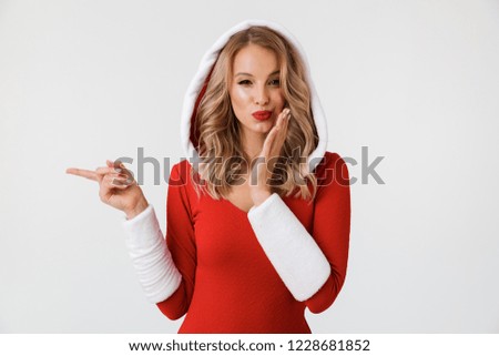 Portrait of a cheerful blonde woman dressed in red New Year costume standing isolated over white background, pointing finger at copy space