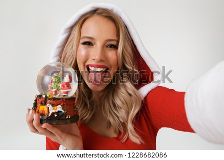 Close up portrait of a funny blonde woman dressed in red New Year costume standing isolated over white background, holding snowball, taking a selfie