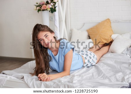 Young girl wearing stylish light blue pajamas  lying down on bed with white and grey sheets decorated with flowers in the light bedroom 