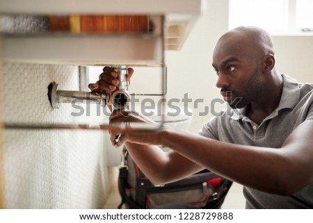 Young black male plumber sitting on the floor fixing a bathroom sink, close up Royalty-Free Stock Photo #1228729888