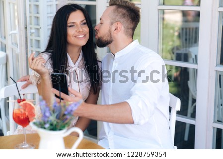 Portrait of lovers, the man takes pictures on the phone as he kisses his beloved. the girl sits with a guy in a cafe, is photographed, smiling and having fun, a guy gently kisses her on the cheek