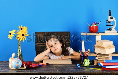 Schoolgirl sits at desk with colorful stationery, books, globe, clock and flowers. Back to school and childhood concept. Kid and school supplies on blue background. Girl with tired face expression