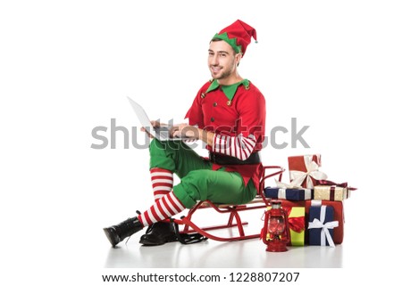 smiling man in christmas elf costume sitting on sleigh near pile of gifts and using laptop isolated on white
